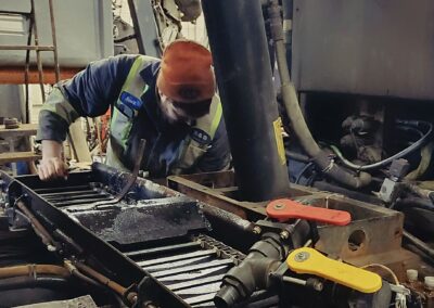 An S&B mechanic in a high-visibility vest and orange toque performing maintenance on heavy equipment