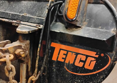 Tenco spreader unit with an amber LED marker light and heavy-duty chains, mounted on a truck inside a maintenance garage.