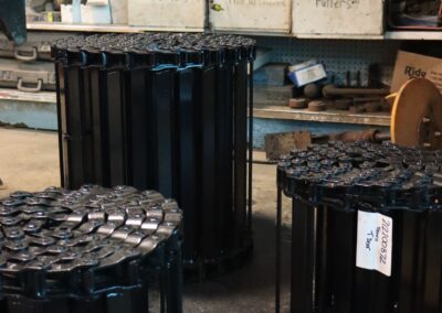 Three freshly painted black roller chain sprocket assemblies standing upright on a parts room floor, with labeled storage shelves in the background.