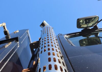 Low-angle upward view of a perforated chrome exhaust stack on a blue heavy-duty truck cab against a clear blue sky.